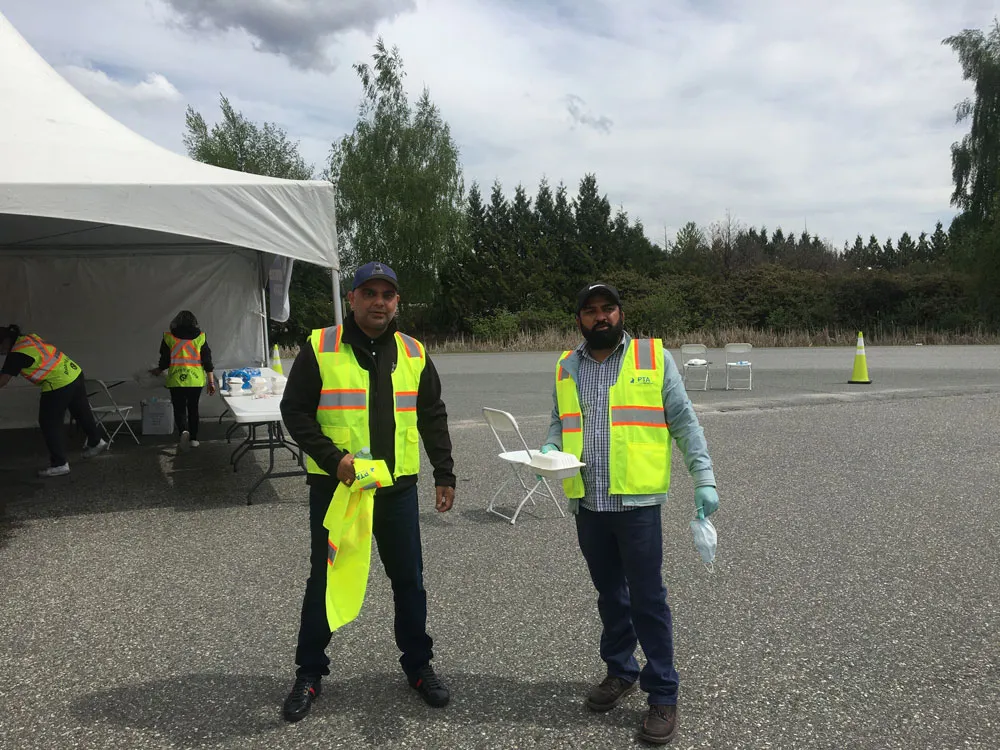 Two volunteers in safety vests by the tent, one holding a takeout meal container ready to hand to a driver
